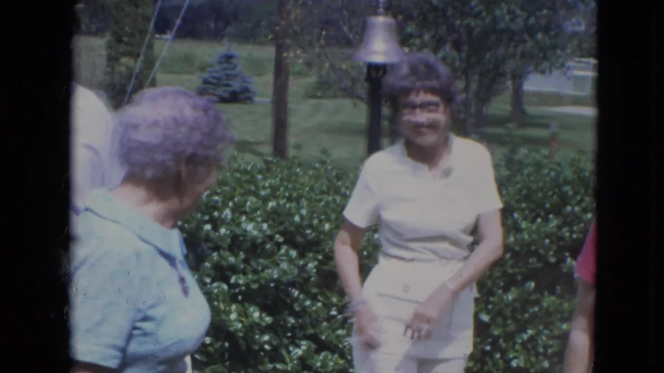 1963 Lincoln, Nebraska home-movie frame of adults gathered in a green garden near a house, one woman smiling toward the camera.