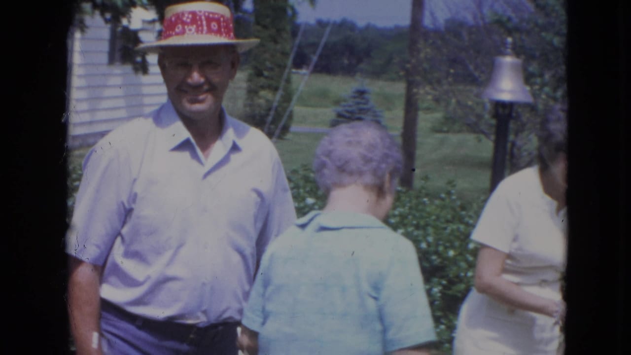 Man in a light shirt and red-banded hat stands near two women in a garden.