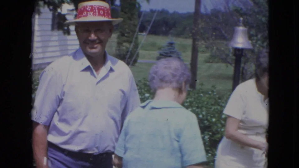 1963 Lincoln, Nebraska frame showing a man in a light blue shirt and red-banded hat with adults gathered in a yard.