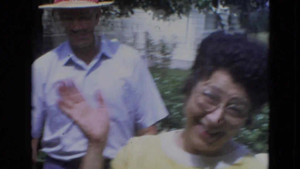 1963 Lincoln, Nebraska frame of a woman in yellow smiling and waving, with a man in a hat behind her in the garden.