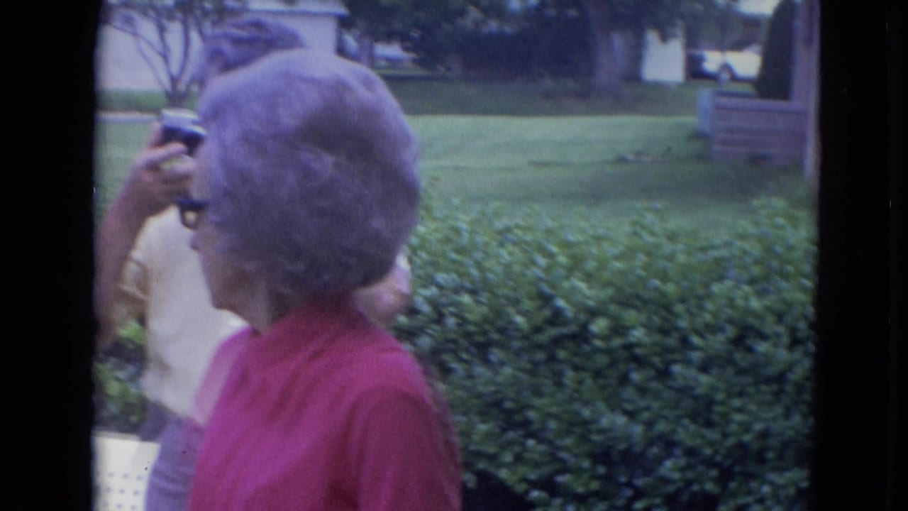 In a 1963 Lincoln, Nebraska yard scene, an older woman in a red shirt turns beside hedges as another adult is partly visible with a camera.