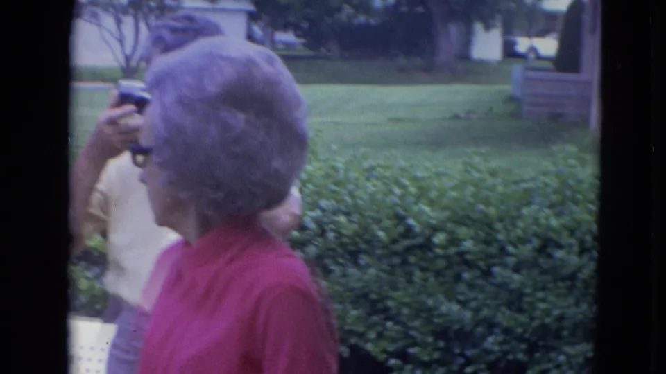 In a 1963 Lincoln, Nebraska yard scene, an older woman in a red shirt turns beside hedges as another adult is partly visible with a camera.