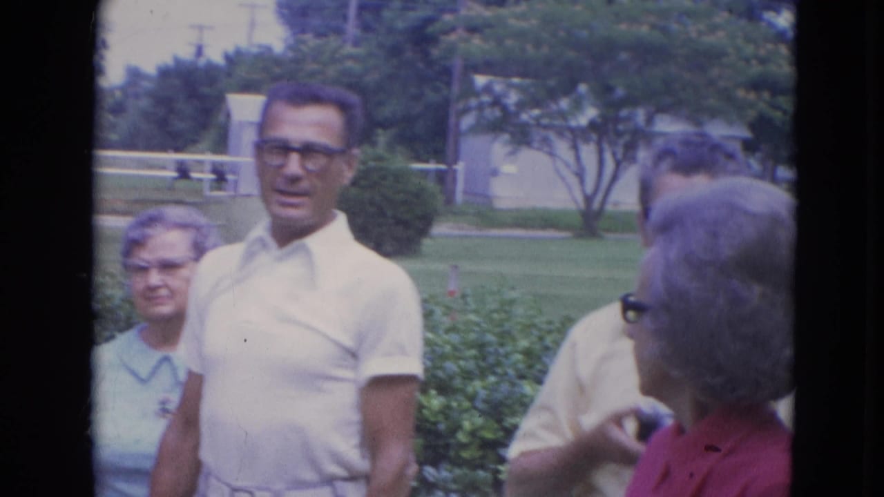 Older adults in a Lincoln, Nebraska suburban yard stand near hedges, including a man in a white polo and glasses and women in light clothing.