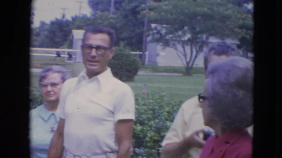 Older adults in a Lincoln, Nebraska suburban yard stand near hedges, including a man in a white polo and glasses and women in light clothing.
