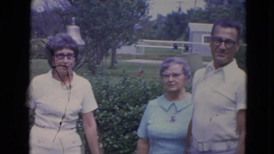 Three older adults pose near a hedge in the 1963 Lincoln yard footage, with two women and a man visible in soft 8mm color.