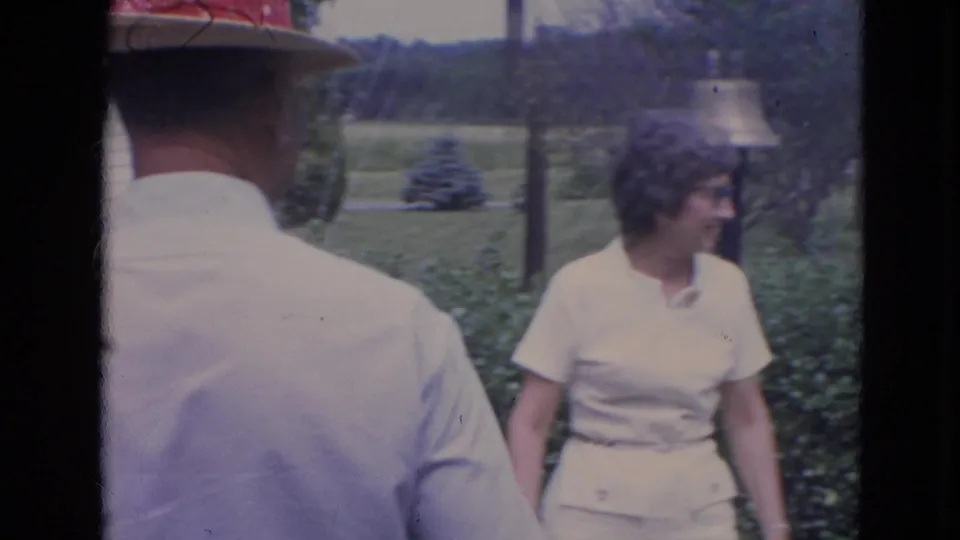 A woman in white turns in the suburban yard as a man in a hat is seen from behind, with hedges, lawn, and distant houses visible.