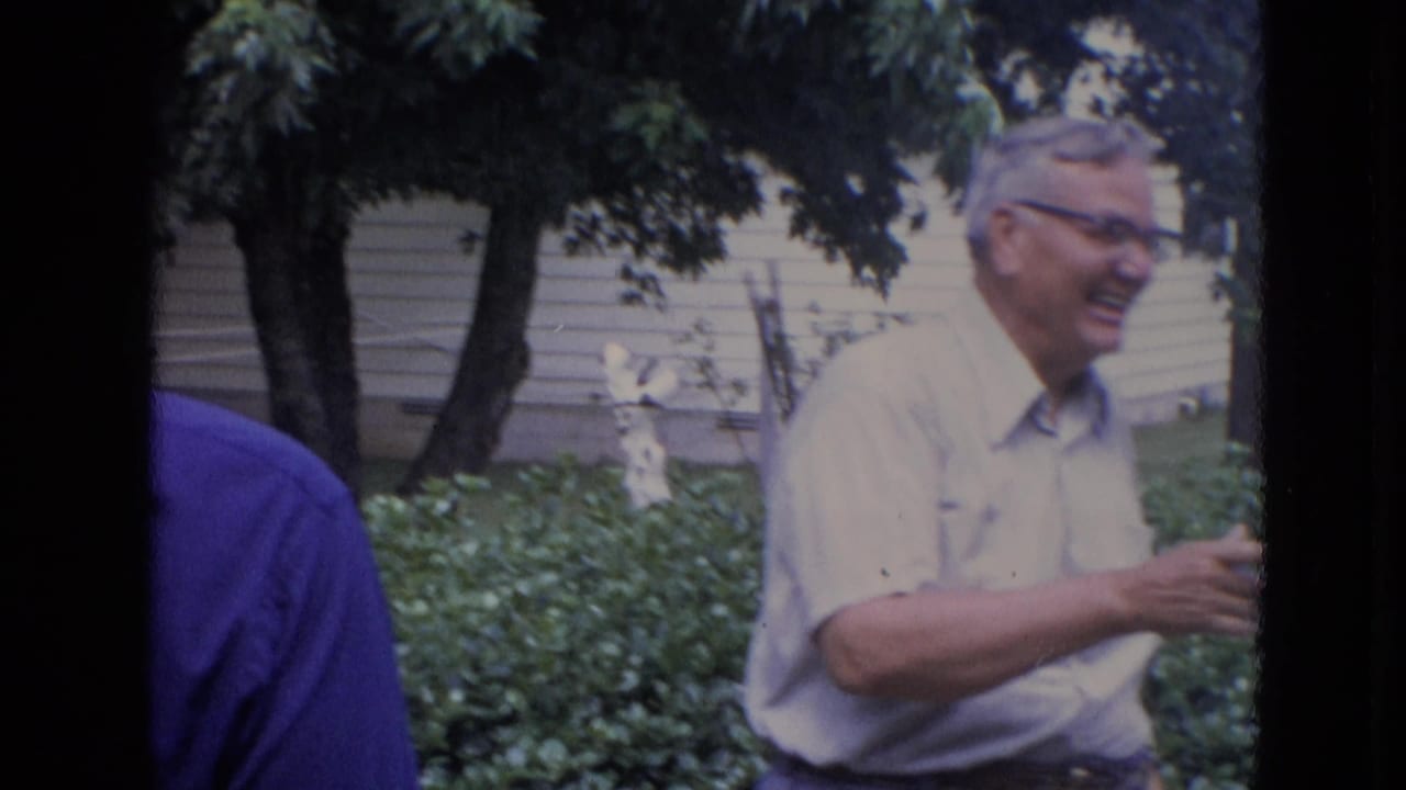 Smiling man with glasses gestures while adults stand in a backyard.