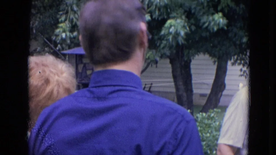 1963 Lincoln, Nebraska home-movie frame showing adults from behind during a casual backyard conversation.