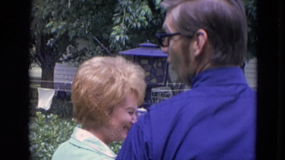 1963 Lincoln, Nebraska frame showing a smiling blonde woman and a man in a blue shirt with glasses in a leafy backyard.