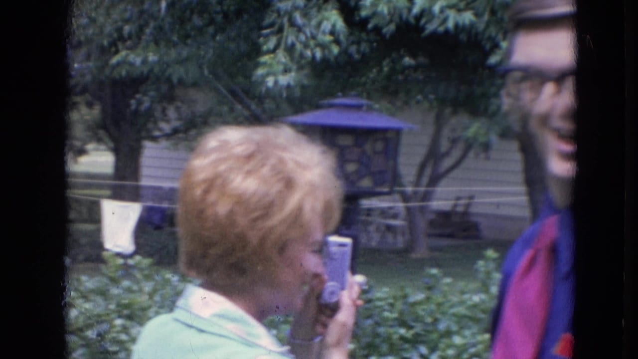 Woman in a light green top holds a small camera while adults stand in a backyard.
