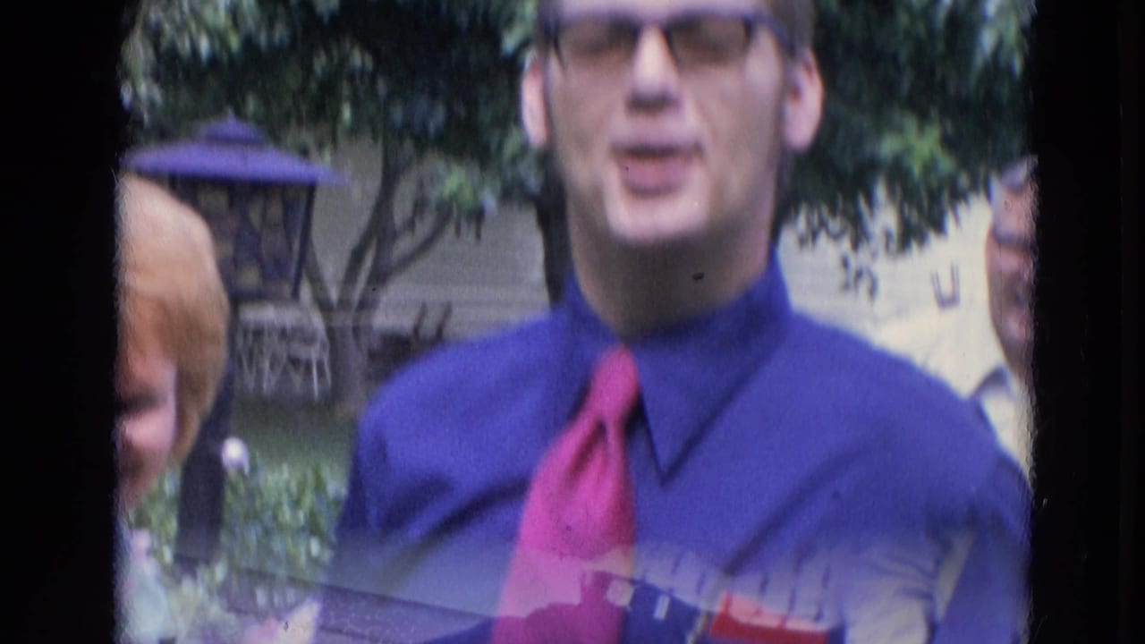 Man in glasses, blue shirt, and red tie stands near a woman in a backyard.
