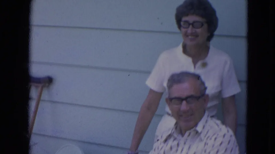 An older man in patterned shirt and glasses poses below a smiling woman against light-colored house siding.