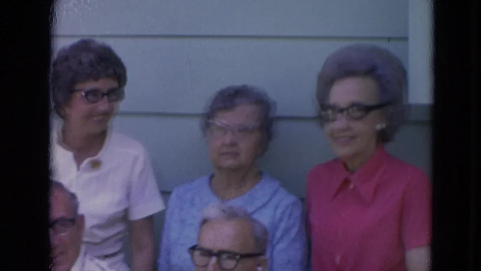 1963 Lincoln, Nebraska home-movie frame showing older adults in glasses gathered outdoors by light house siding.