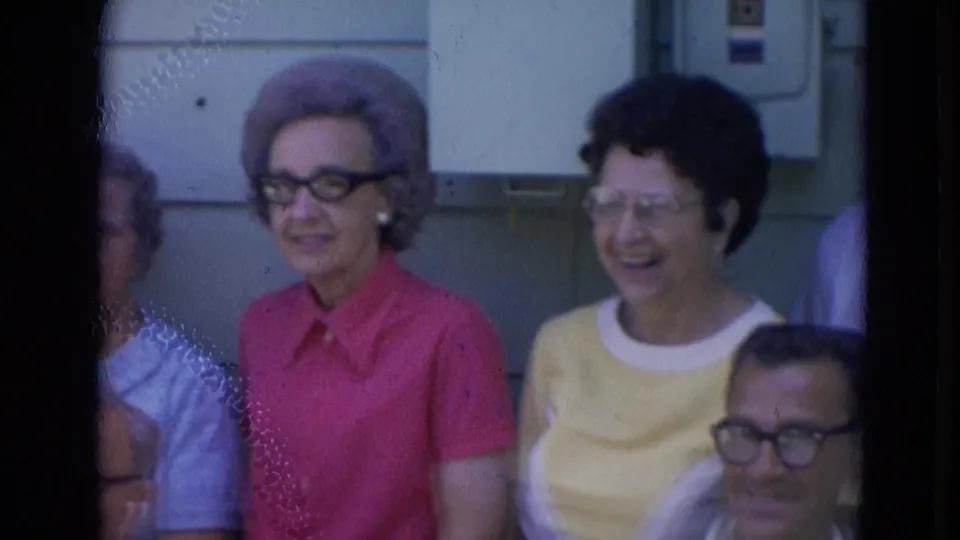 1963 Lincoln, Nebraska frame with smiling older women in pink and yellow tops beside seated men outdoors.