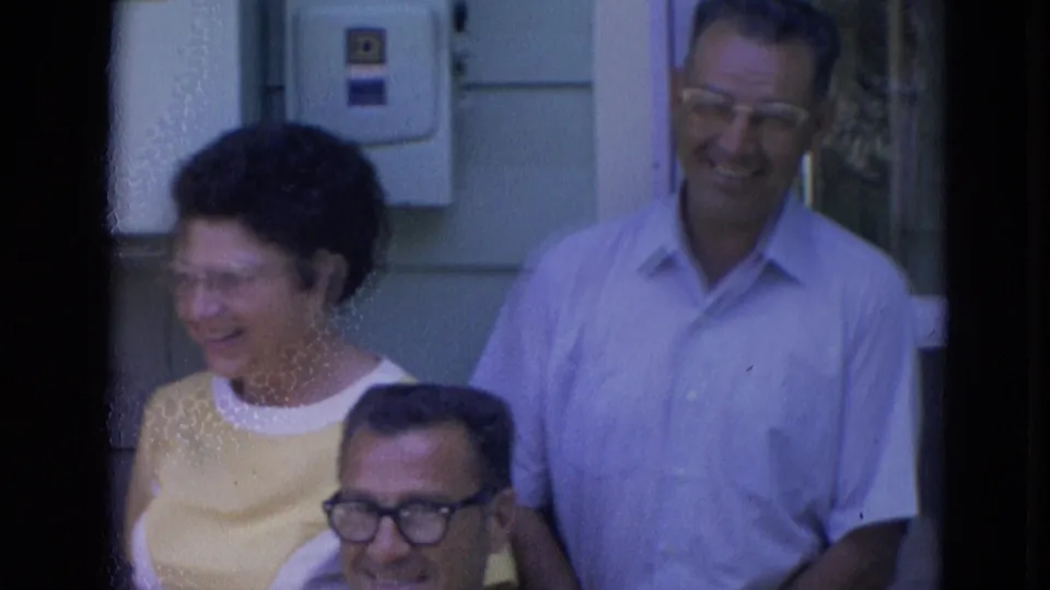 1963 Lincoln, Nebraska home-movie frame showing a woman laughing near a standing man and seated man outdoors.