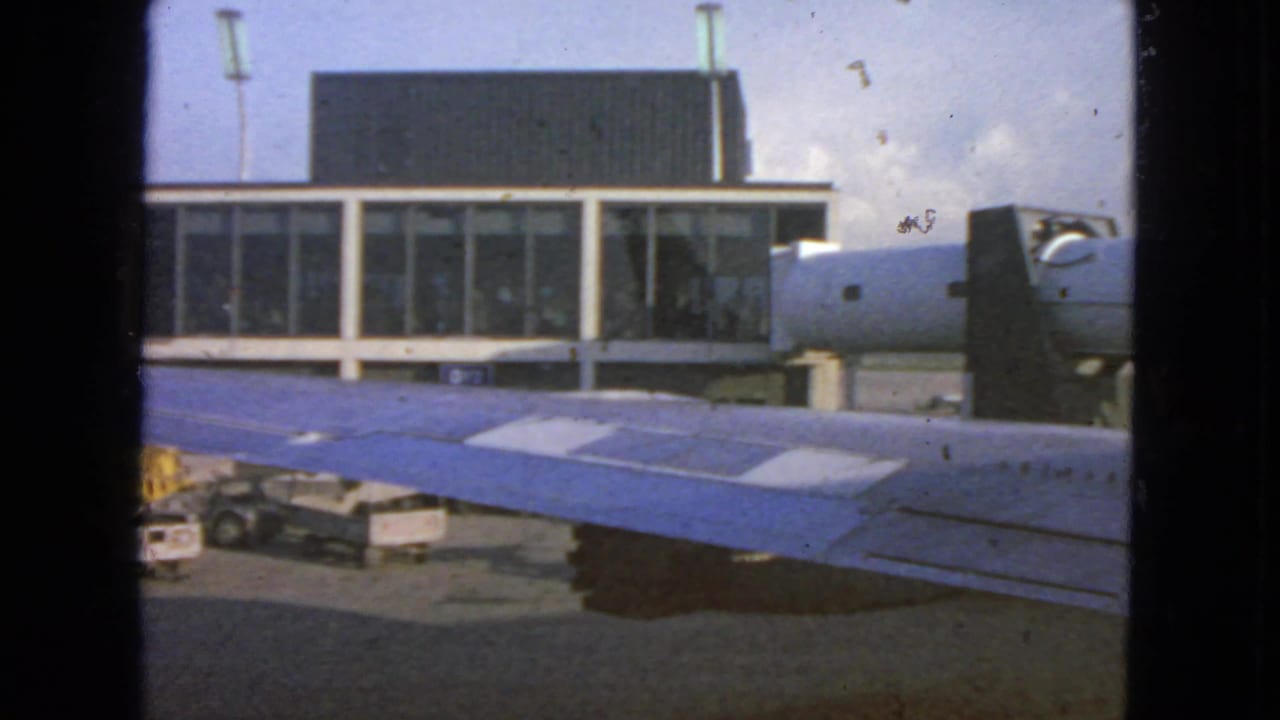 1963 San Francisco airport view from inside an aircraft, showing a blue wing, terminal windows, ground carts, and bright sky.