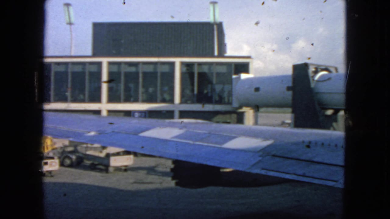 Airport terminal seen across a blue airplane wing with service carts below.