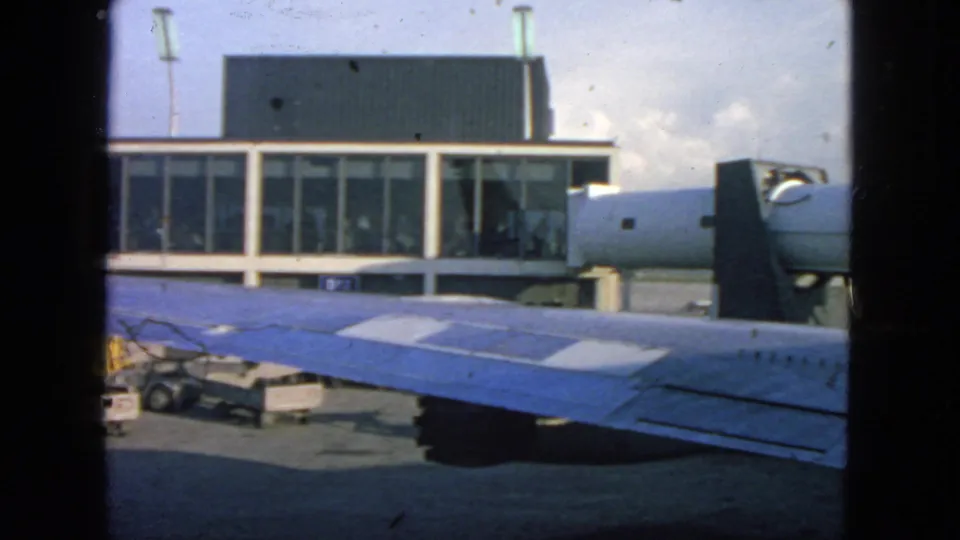 1963 San Francisco airport scene from a passenger window, with a blue wing, terminal facade, ground carts, and small figures behind glass.