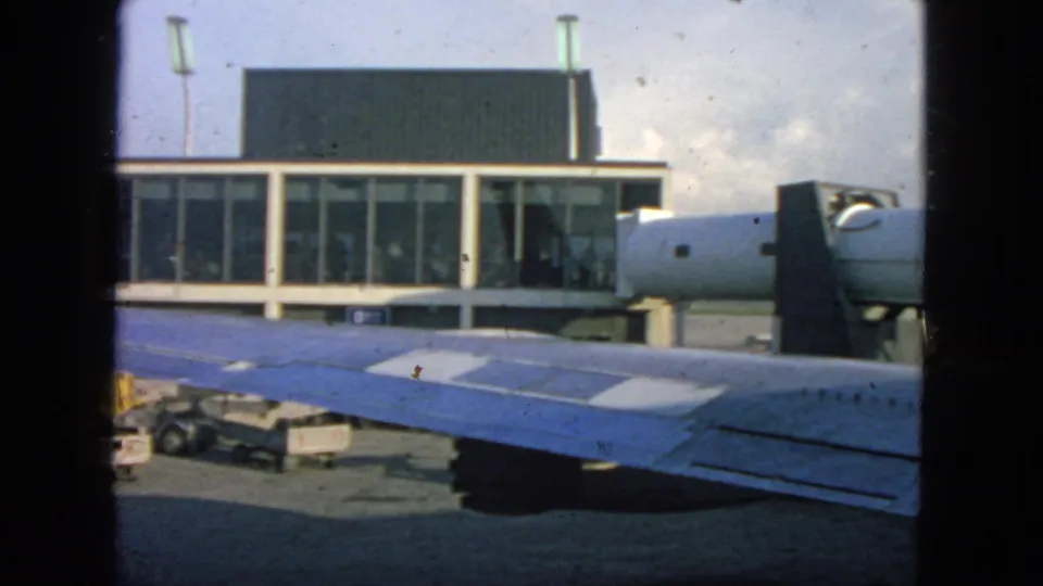 1963 San Francisco airport gate view with a blue aircraft wing in front, terminal windows, service carts, and indistinct people inside.