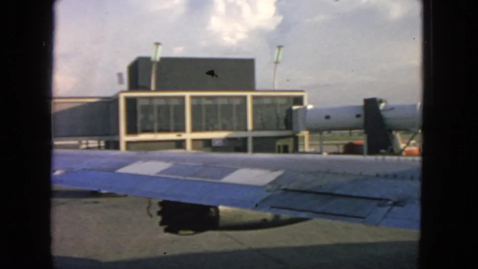 1963 airplane-window view with wing and engine area below a glass-fronted San Francisco airport terminal.