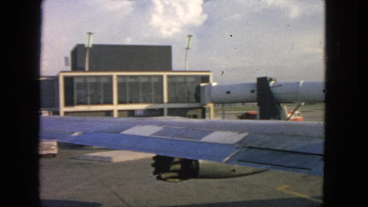 Airplane wing seen through a window with terminal building and jet bridge beyond.