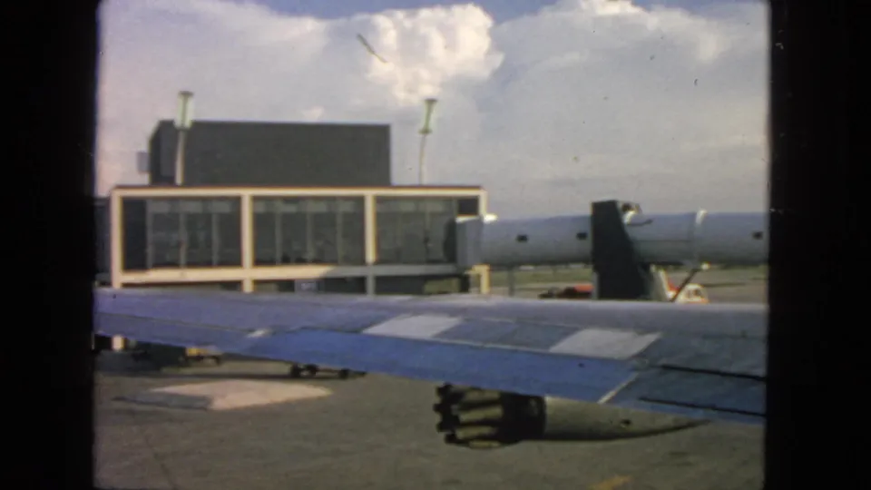 1963 airplane-window frame showing a blue-and-white wing, engine, modernist terminal facade, and partly cloudy sky in San Francisco.