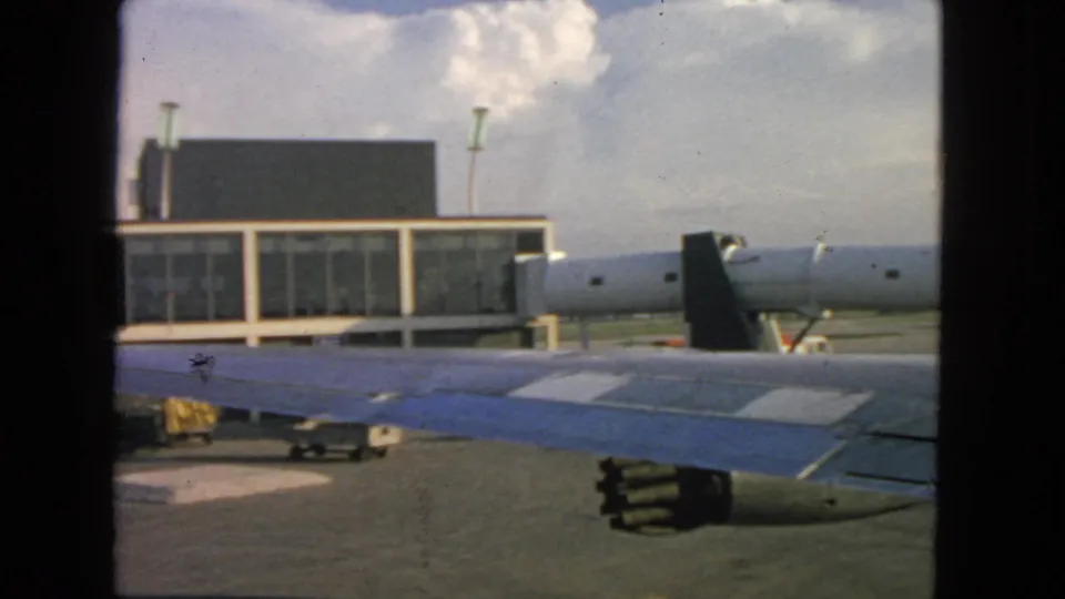 1963 San Francisco airport tarmac view with aircraft wing, engine, terminal glazing, jet bridge, and a small ground-service cart.