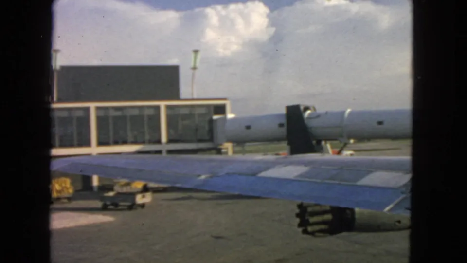 1963 passenger-window view at San Francisco showing an aircraft wing crossing the frame before terminal windows and a jet bridge.