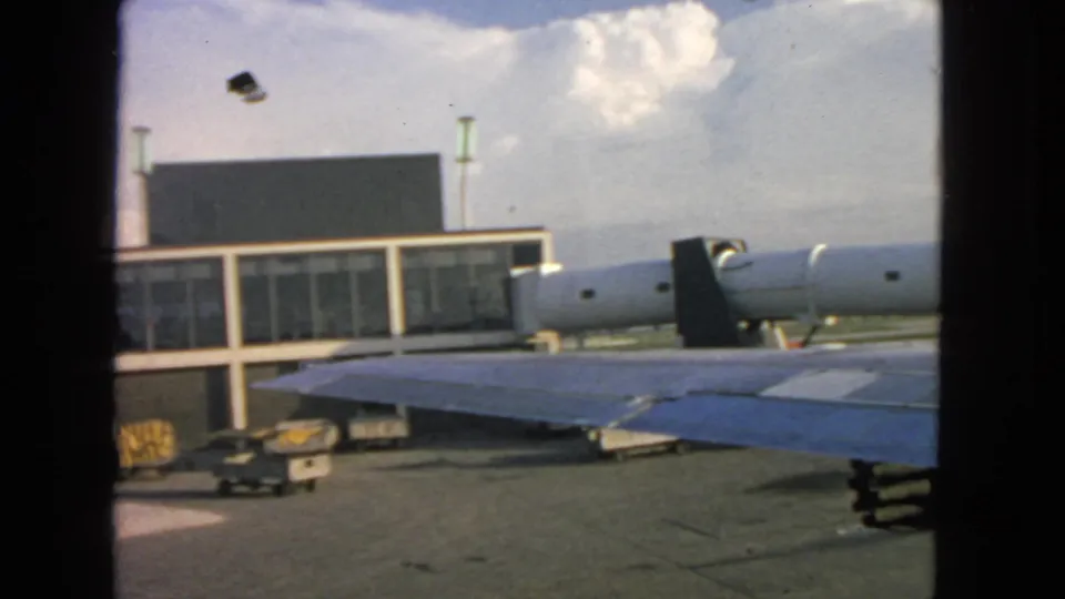 Aircraft-window view at San Francisco airport in 1963, showing a blue wing, engine nacelle, jet bridge, terminal facade, and baggage carts.