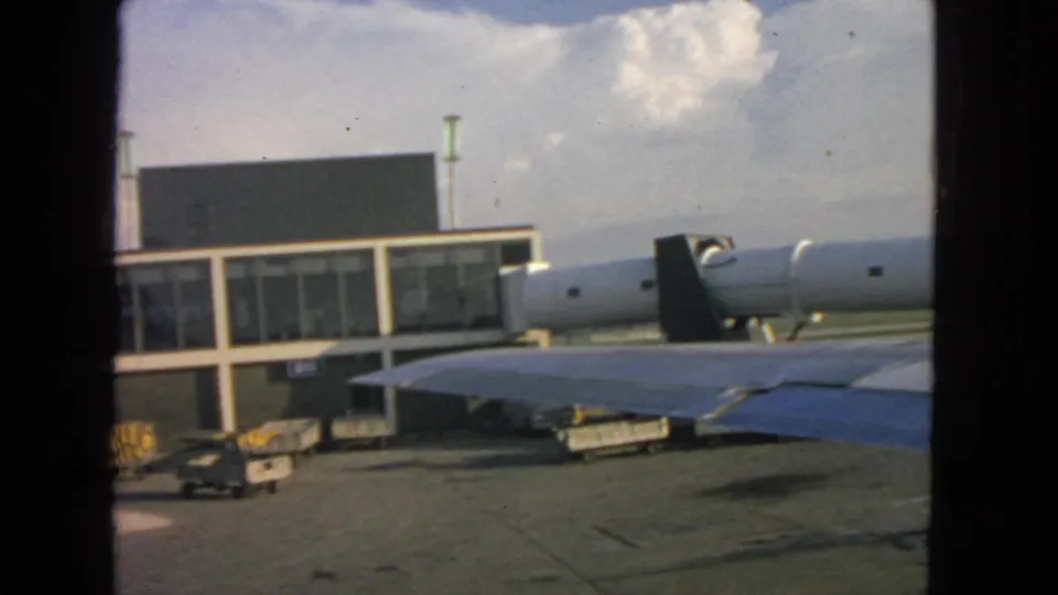 1963 home-movie frame from a passenger aircraft window, with the wing crossing the tarmac view toward the terminal and distant landscape.