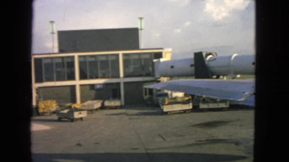 Passenger-window view at San Francisco airport in 1963, looking over the aircraft wing toward terminal windows, jet bridge, carts, and open sky.