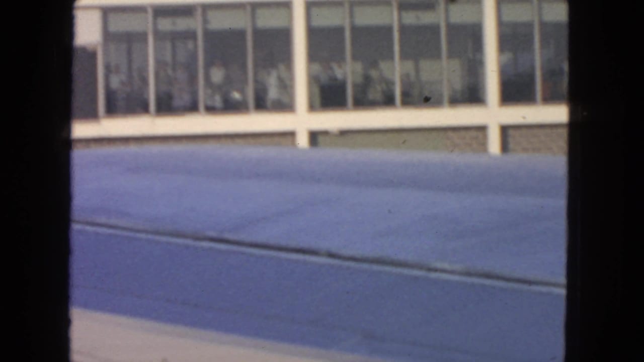 Aircraft wing fills the lower frame while a 1963 San Francisco airport terminal with people behind windows appears beyond it.