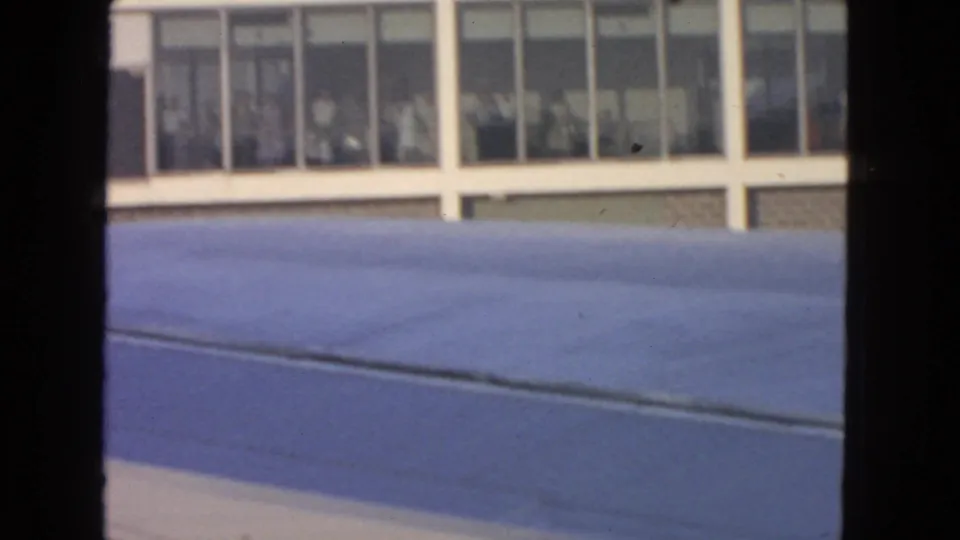 Aircraft wing fills the lower frame while a 1963 San Francisco airport terminal with people behind windows appears beyond it.
