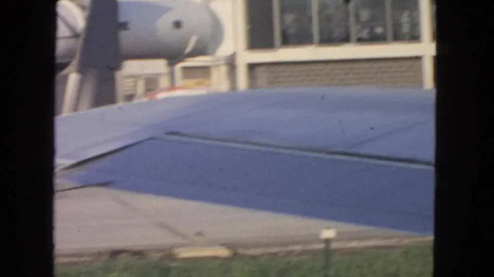 Blue aircraft wing dominates the foreground near a San Francisco airport terminal and boarding equipment in 1963 footage.