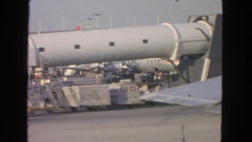 1963 San Francisco airport tarmac view with an aircraft wing, cylindrical jet bridge, service vehicles, and terminal structures.