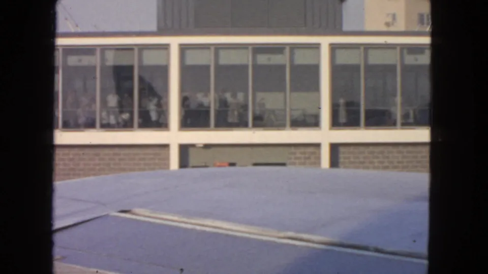 1963 airport terminal facade seen over an aircraft wing, with several people faintly visible inside the windowed building.