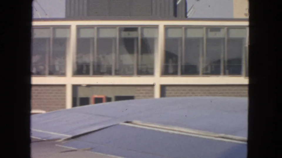 San Francisco airport terminal windows in 1963 seen from near an aircraft wing, with faint figures inside and boarding areas nearby.
