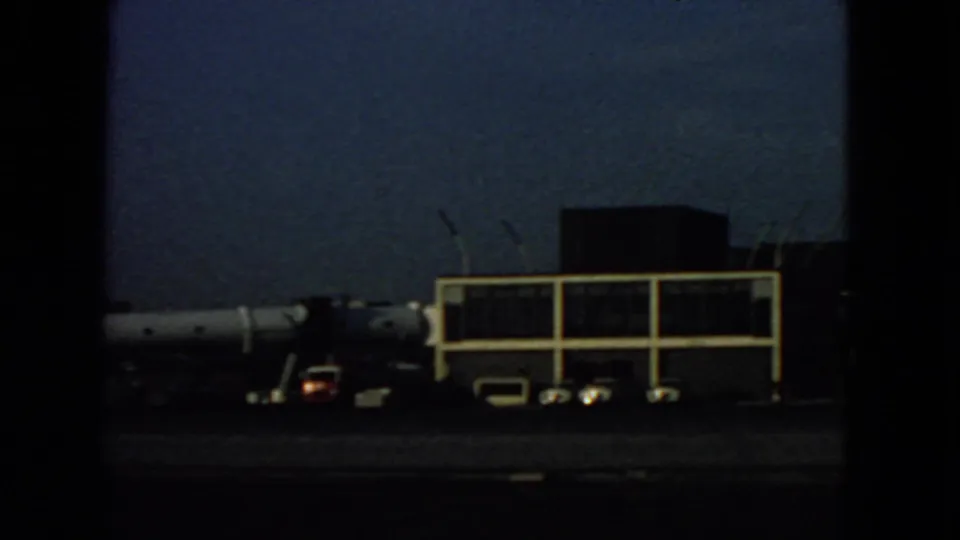 Dim 1963 airport tarmac view from inside a plane, with a terminal structure, parked aircraft, vehicles, and blue sky.