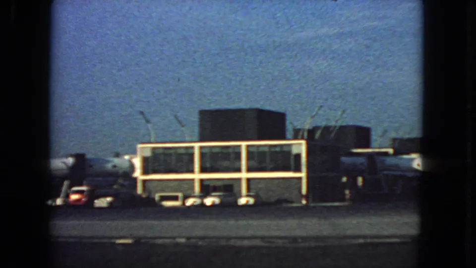 1963 San Francisco airport tarmac scene with a glass-fronted terminal, aircraft, ground vehicles, and handheld film grain.