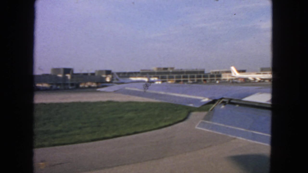 Airplane wing and flaps seen through a window with terminal buildings and parked jets beyond.