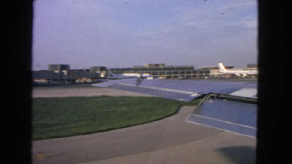 1963 San Francisco airport view from an airplane window, showing the wing, flaps, tarmac, terminal buildings, and parked aircraft.