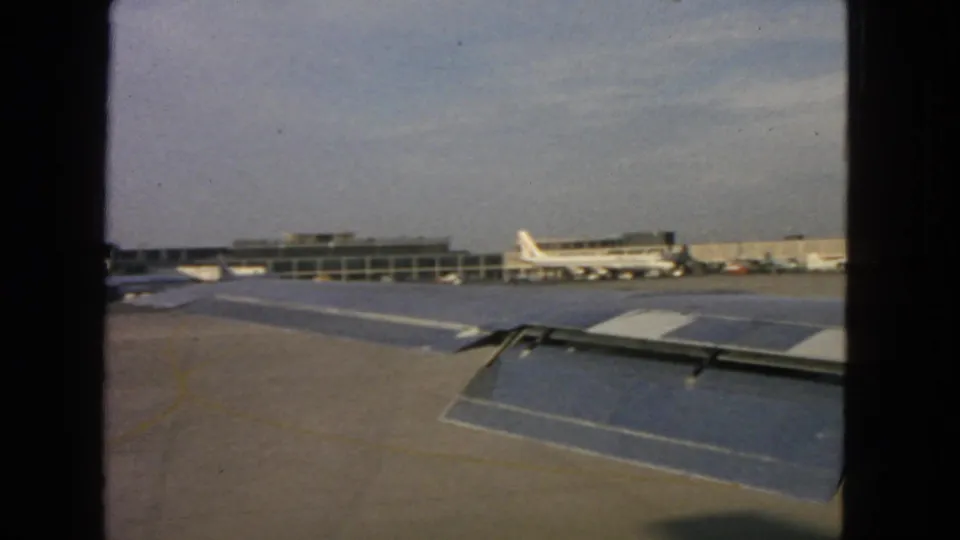 San Francisco airport in 1963 seen from an airplane window, with wing flaps, terminal gates, parked jets, and open tarmac visible.