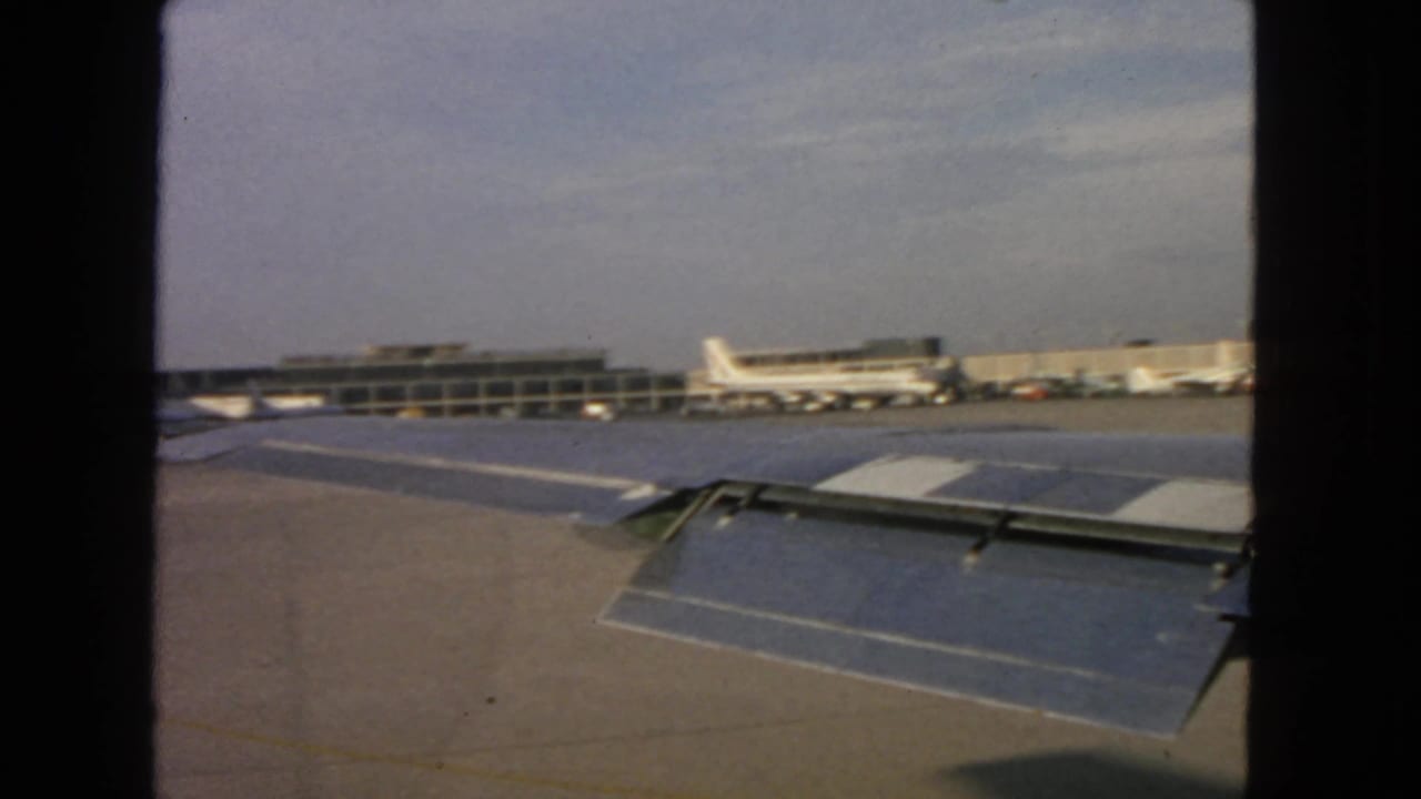 1963 passenger-window view at San Francisco International Airport showing an airplane wing, tarmac, terminal, and parked aircraft.