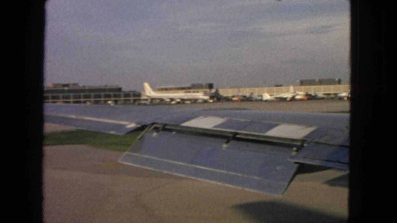 Aircraft wing over the tarmac with terminal buildings and parked planes in the distance.