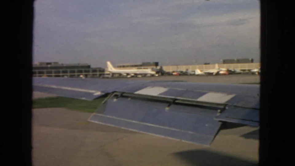 Passenger view over an aircraft wing at the San Francisco airport tarmac, with low terminal buildings and distant parked planes.
