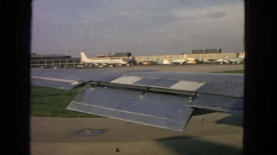 1963 home-movie frame from an aircraft window showing a wing, grass strip, tarmac, and distant terminal area at San Francisco airport.