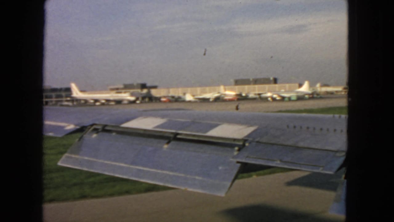 1963 San Francisco airport view from an airplane window, showing wing flaps, parked aircraft, terminal buildings, and tarmac.