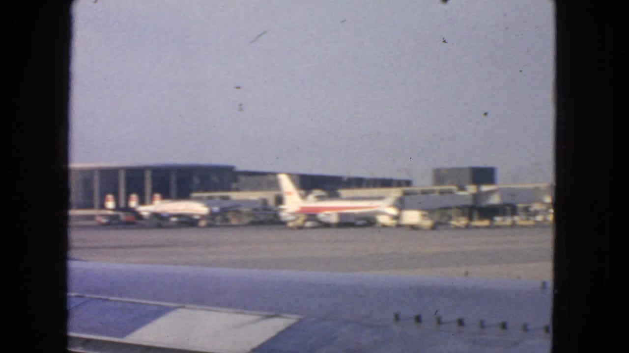Tarmac view with airplane wing edge, jetliners, terminal structures, and ground equipment.