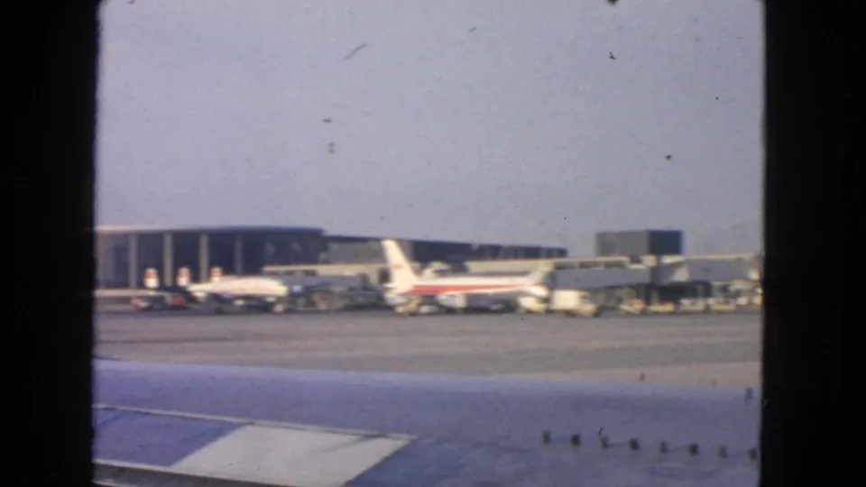 1963 San Francisco airport tarmac seen from inside an aircraft, with parked jetliners, gate equipment, and terminal structures.