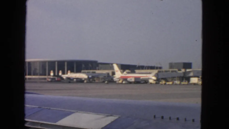 1963 San Francisco airport view across an airplane wing toward parked aircraft, terminal buildings, and ground service equipment.